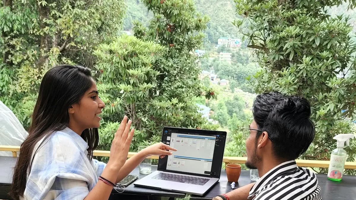 Two young Indian students talking over a laptop at an outdoor table with green hills behind them