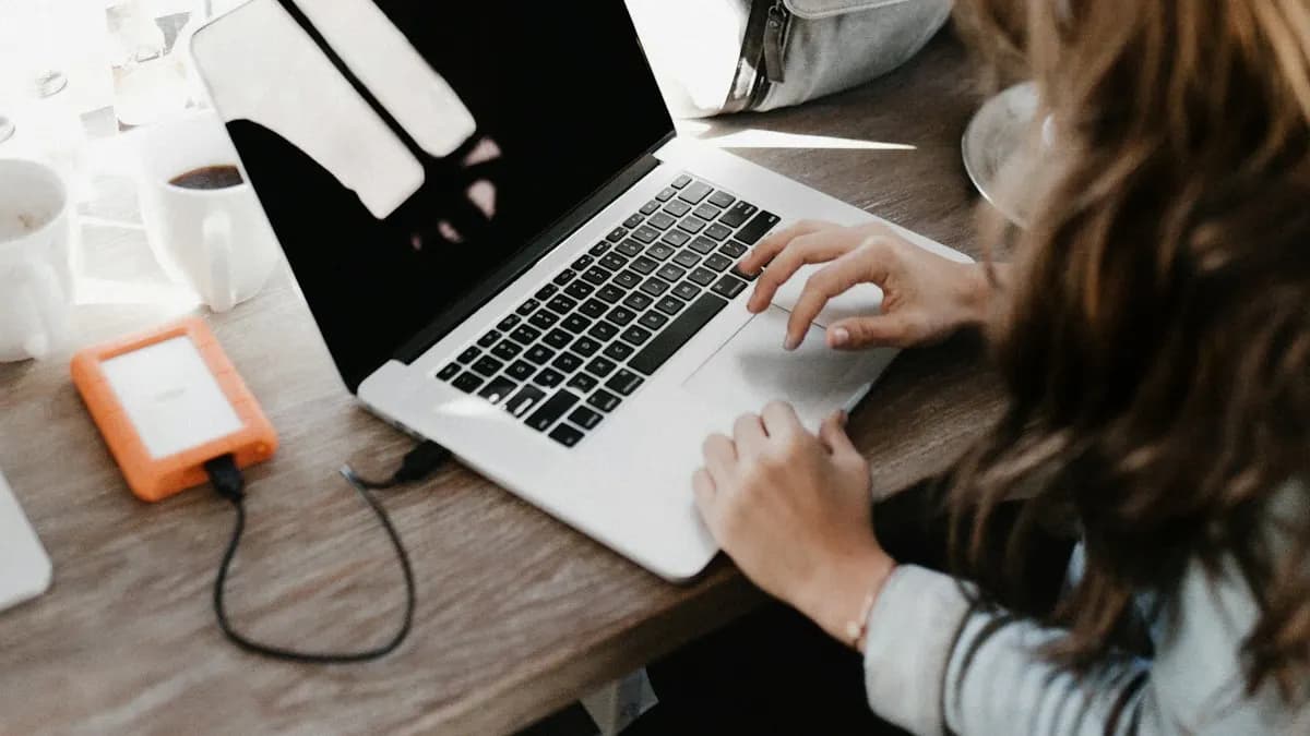 College student typing on a laptop at a wooden table with a portable hard drive beside it