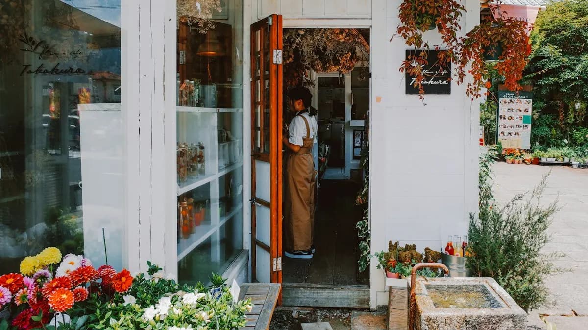 Small flower shop entrance with plants and fresh flowers outside and the owner standing in the doorway