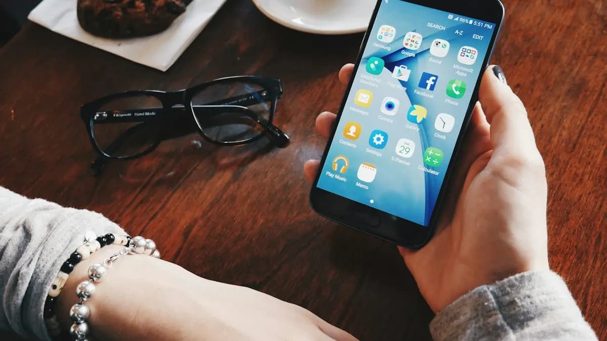 Hand holding a smartphone with apps on screen over a wooden table with glasses