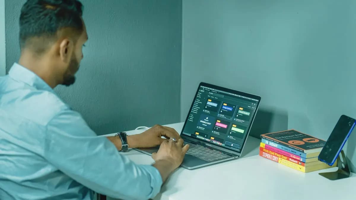 Young designer working on a laptop at a bright desk with a project board on screen and books beside him