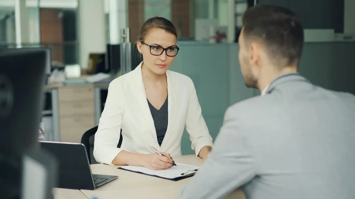 Young woman in a white blazer interviewing a candidate across an office desk with a laptop