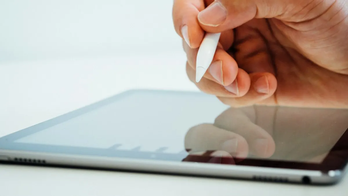 Hand holding a white stylus above a tablet screen on a bright desk