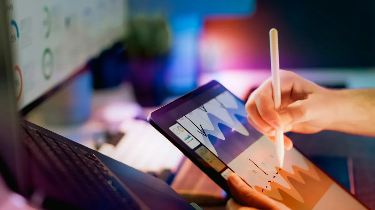 Hand using a stylus on a tablet showing sales charts beside a laptop at night